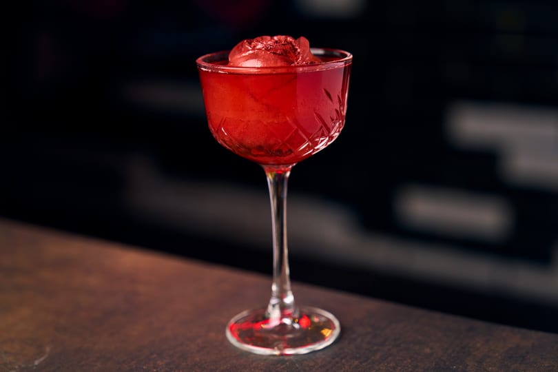 Red cocktail in a coupe glass with a rose-shaped ice cube on a dark bar.