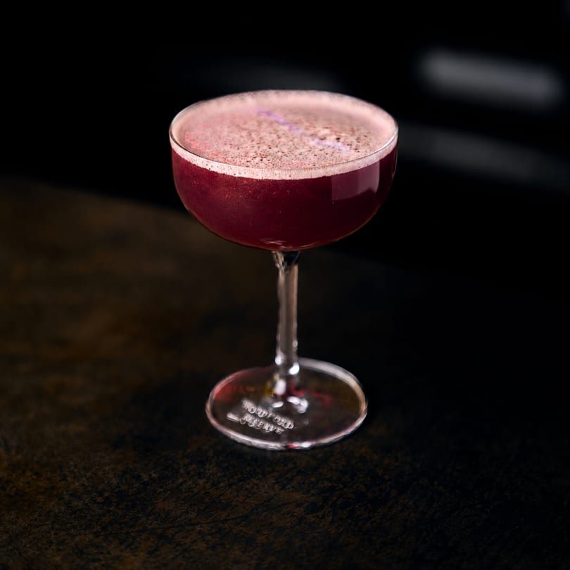 Dark red foamy cocktail in a Woodford Reserve coupe glass on a dark surface.