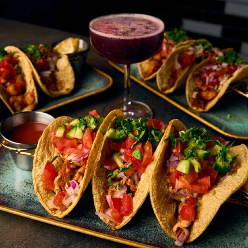 Three tacos with red salsa, avocado, and cilantro, with a purple cocktail and more tacos in the background.