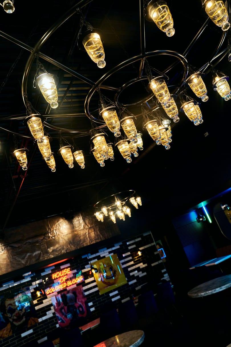 Ornate chandelier with warm glowing lights illuminates a dark restaurant interior with a neon sign and tiled wall.