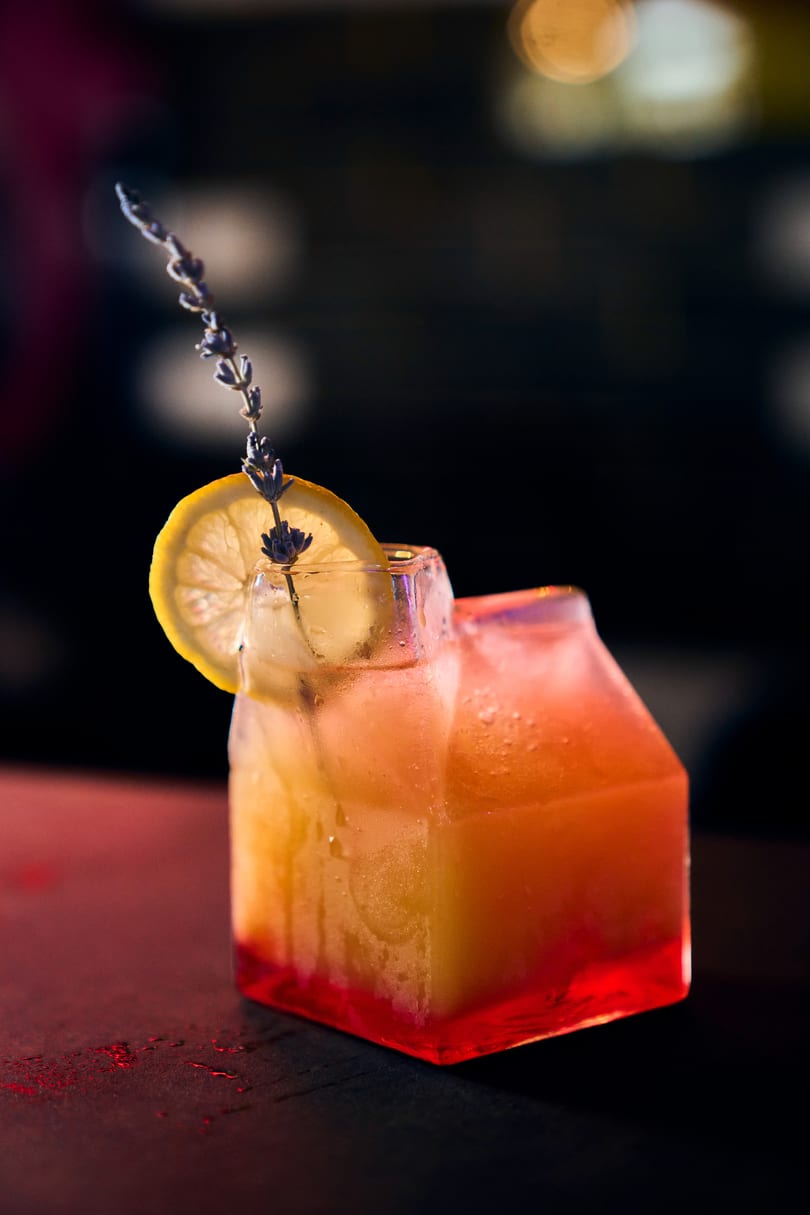 Orange and red cocktail in a square glass, garnished with lemon and lavender, on a dark bar.